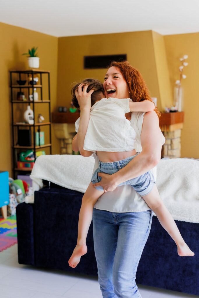 Mère riant aux éclats en portant son enfant dans un salon chaleureux — photographe famille Cergy