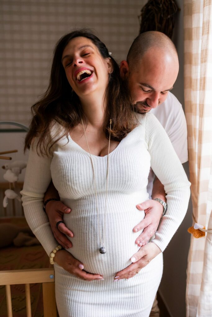 Couple complice devant le berceau, future maman riant aux éclats, mains sur le ventre — photographe grossesse Cergy