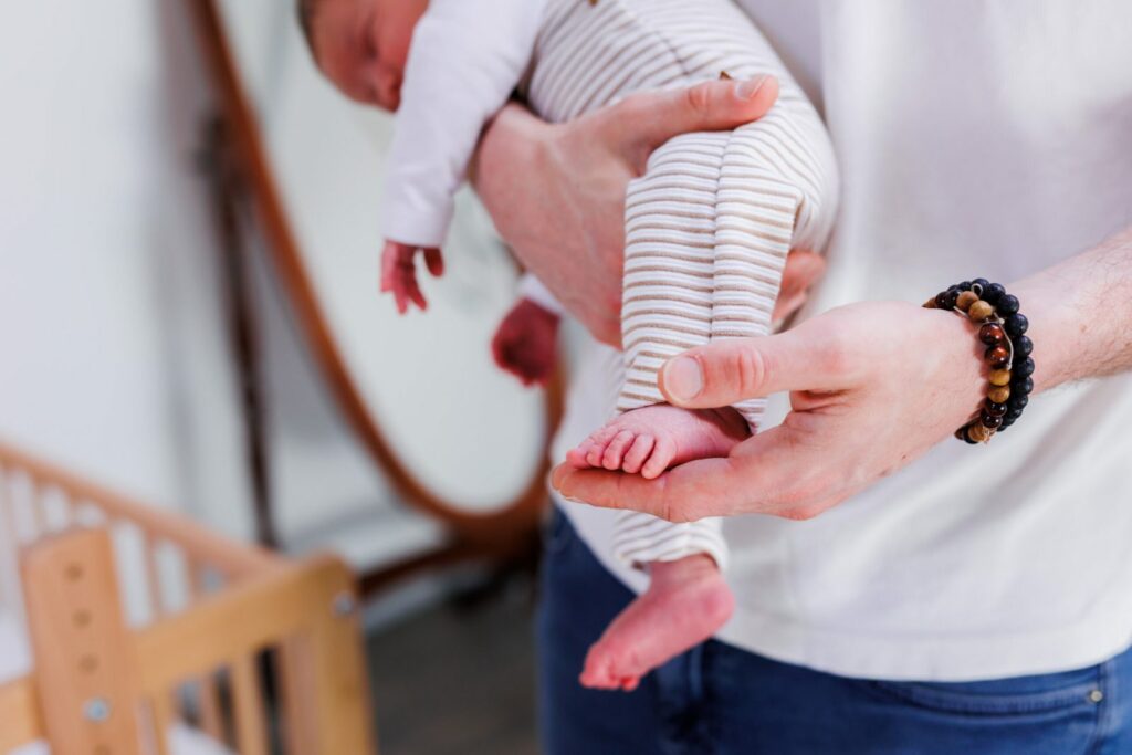 Papa portant son bébé, petit pied niché dans sa main, chambre avec berceau — photographe naissance Cergy