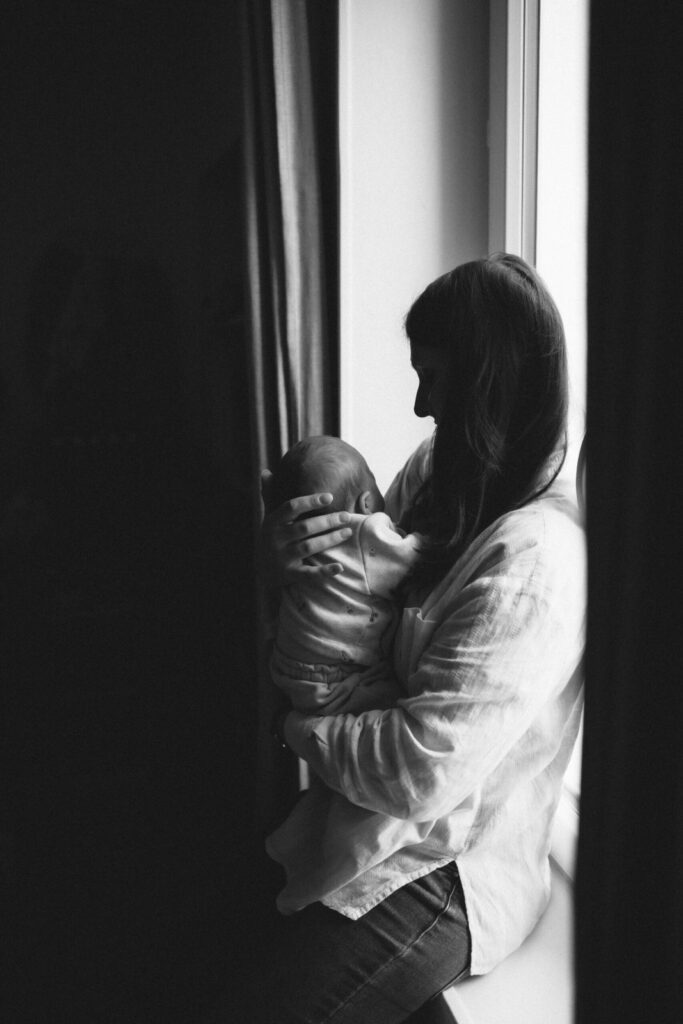 Maman assise devant la fenêtre, berçant tendrement son bébé contre elle, noir et blanc