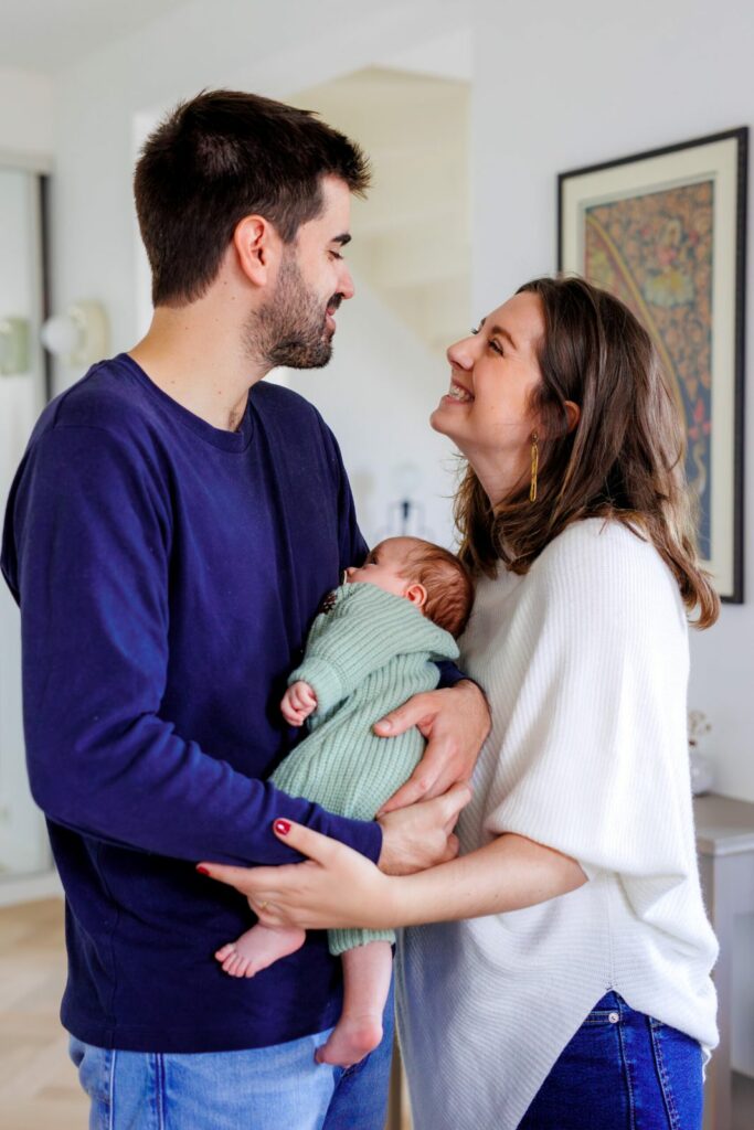 Les parents riant ensemble, tenant leur bébé endormi en pyjama vert, salon lumineux